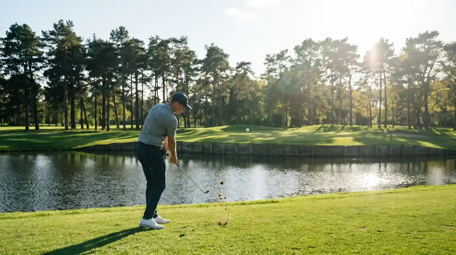 Professional golfer playing a precise iron approach to an elevated green guarded by a fronting pond on a parkland course