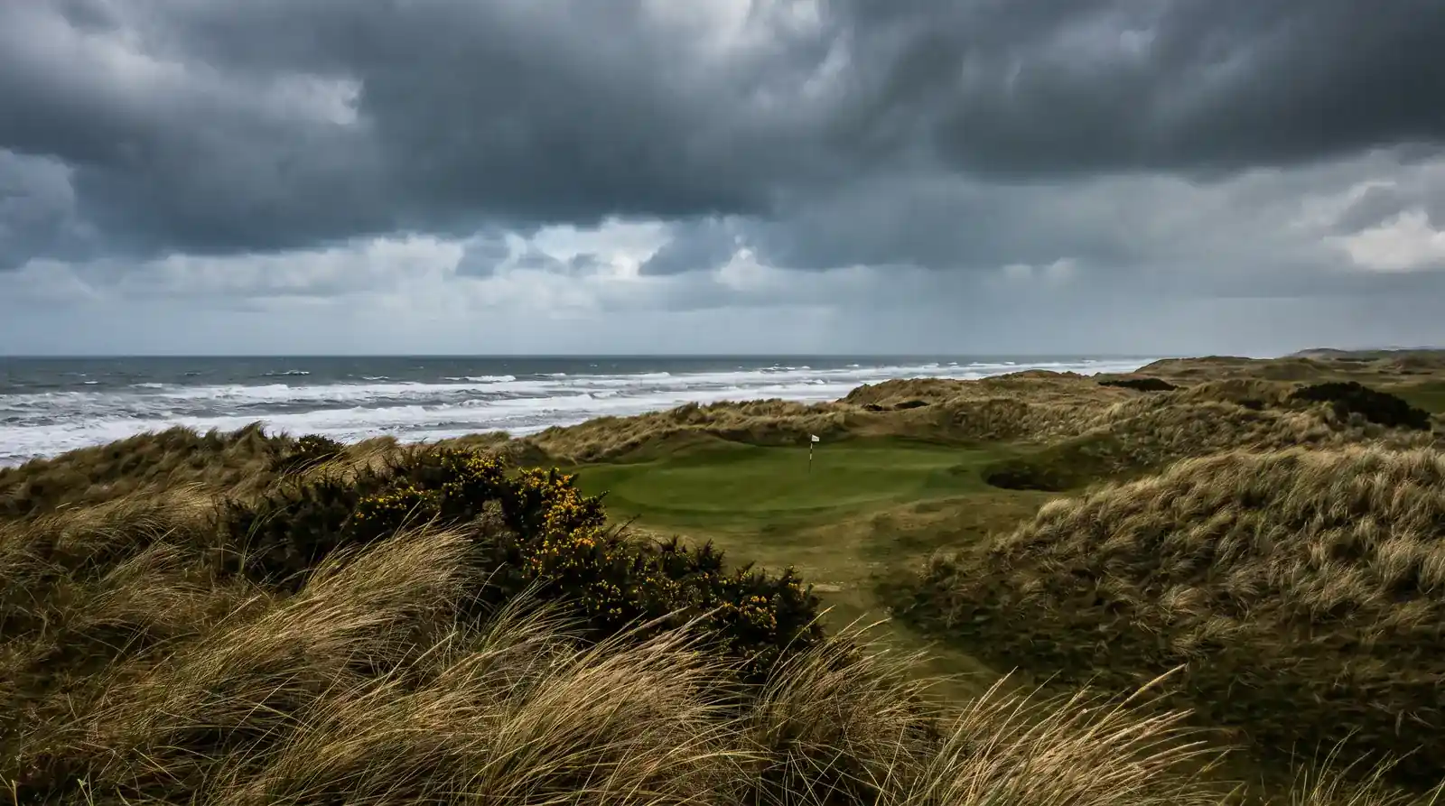 Windswept links course with dramatic dark clouds gathering over the coastline behind a tucked-back green